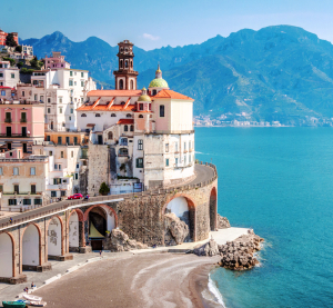 K&uuml;stenstadt Amalfi in Italien mit bunten H&auml;usern auf einer Klippe, darunter eine Strandbucht und das t&uuml;rkisfarbene Mittelmeer, im Hintergrund Berge.