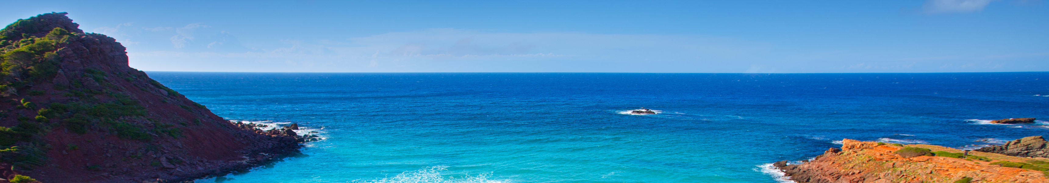 Unber&uuml;hrter Strand Cala Pregonda auf Menorca mit goldenem Sand, t&uuml;rkisblauem Meer und r&ouml;tlichen Felsen unter blauem Himmel