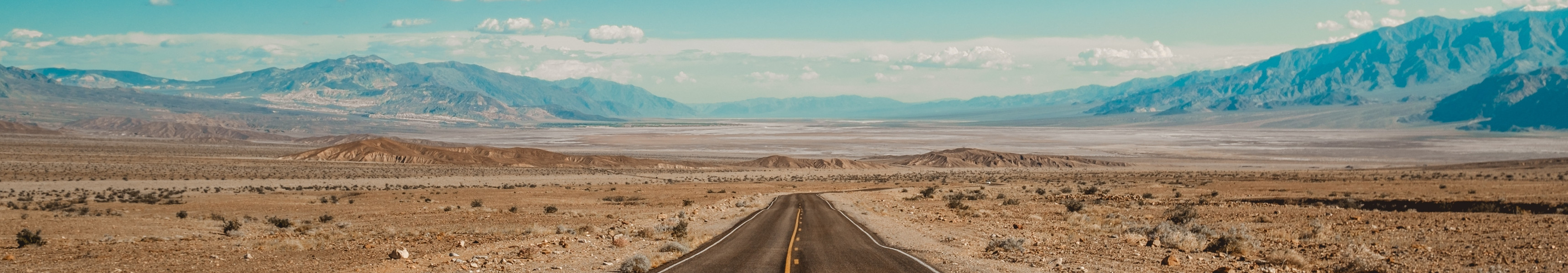 Eine endlos wirkende Stra&szlig;e f&uuml;hrt durch die karge W&uuml;stenlandschaft des Death Valley in Kalifornien, USA, mit Bergen am Horizont unter einem klaren blauen Himmel.