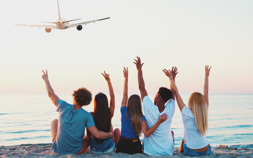 F&uuml;nf junge Erwachsene sitzen nebeneinander im Sand, schauen auf das Meer und heben freudig die Arme in die Luft.