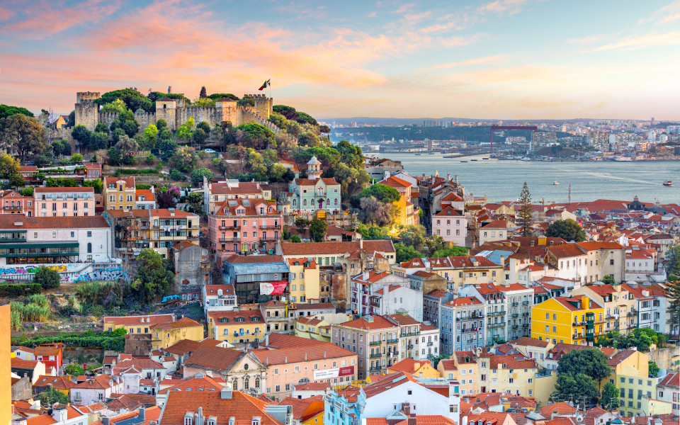 Panoramablick &uuml;ber Lissabon mit der beeindruckenden Burg S&atilde;o Jorge, farbenfrohen H&auml;usern und dem glitzernden Tejo-Fluss im Abendlicht.
