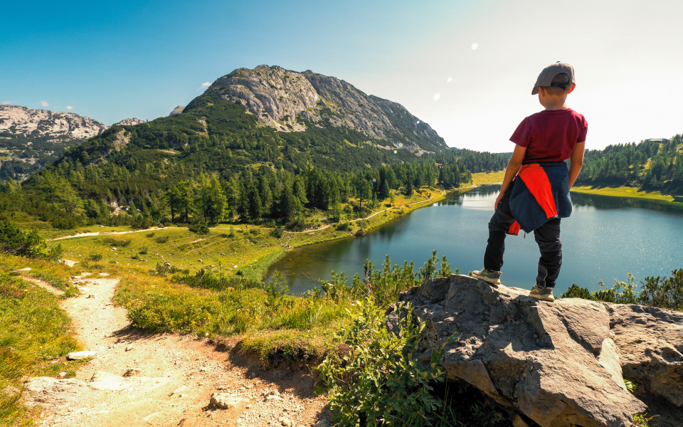 Kind mit Basecap steht auf Felsvorsprung vor einem Bergsee, rundum Almwiesen, Nadelwald und felsige Gipfel.