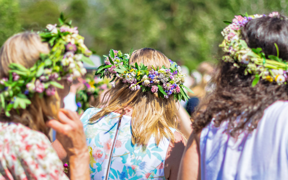 Mehrere Frauen mit Blumenkr&auml;nzen auf dem Kopf feiern im Freien bei sonnigem Wetter das Midsommar Fest.